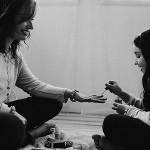Two women sitting on the floor, one painting the other's nails, with nail polish bottles around them.