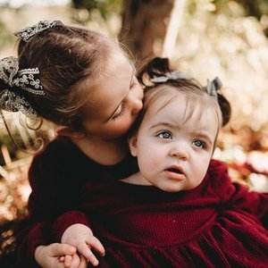 Two young girls sitting outdoors, one toddler with a bow in her hair and the other girl kissing her on the cheek.