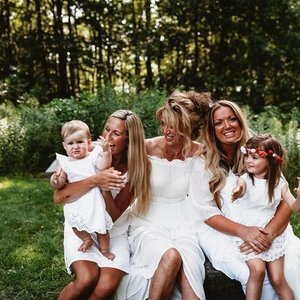 Four women and two young girls sitting outdoors on grass surrounded by trees, all smiling and wearing white dresses.