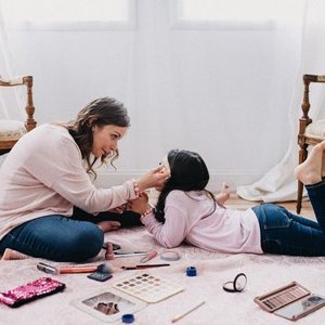 A woman and a young girl are playing on the floor with makeup and beauty products, smiling and having fun.