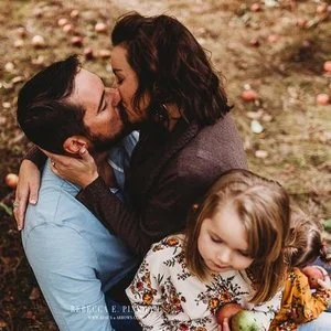 A couple embracing and about to kiss outdoors, with a young girl sitting in front of them holding a pumpkin.