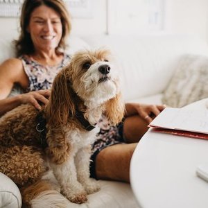 A woman smiling while sitting on a couch with a brown and white dog on her lap, next to an open notebook and a pen.