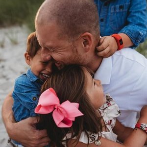 A man hugging two young children, one boy and one girl, outdoors with smiles and affection.