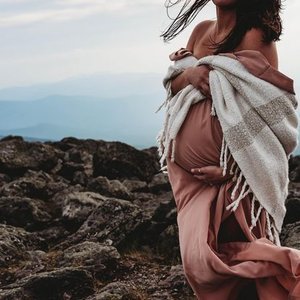 A pregnant woman standing on rocky terrain near a body of water, wrapped in a blanket, with her hands on her belly.