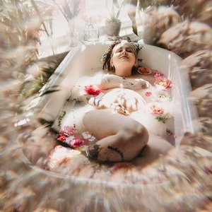 A woman lying in a bathtub filled with water and flower petals, with a peaceful expression, surrounded by blurred pink and beige flowers.