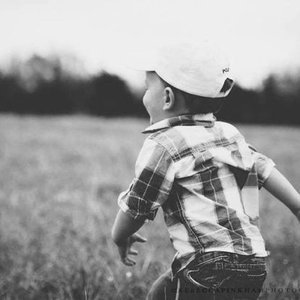 A boy in a baseball cap and plaid shirt sitting outdoors in a grassy field.