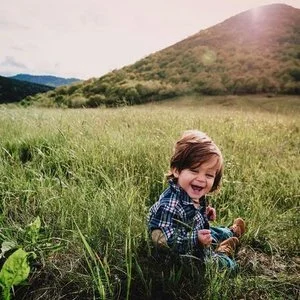 A young boy sitting in tall grass outdoors, smiling and laughing with mountains in the background.