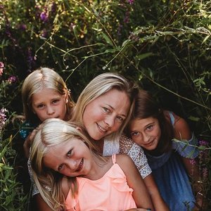 A woman with three young girls outdoors in front of foliage, smiling and close together.