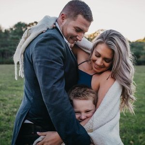 A happy family of three, with a man, woman, and young boy, hugging outdoors in a grassy field during the daytime.