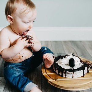 Young child sitting on the floor, eating a chocolate cake with white frosting and Oreos, with a plate of cake nearby.