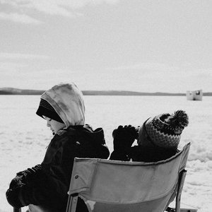 Two children sitting in a chair outdoors near a body of water, dressed in winter clothing, looking down.