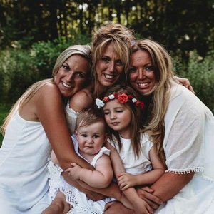 Four women and two young girls smiling outdoors in a garden, dressed in white, with happy expressions.