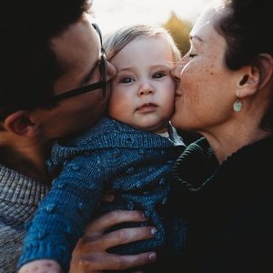 A young child being kissed on both cheeks by two adults, one on each side, outdoors during sunset.