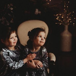 Two young girls wearing matching silver dresses sitting together in a cozy, dimly lit room with a large beige armchair and a decorative vase with string lights in the background.