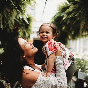 A woman lifting a young girl in a greenhouse filled with hanging plants, both smiling and enjoying each other's company.