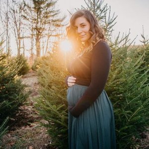 A young woman in a black long-sleeve top and blue skirt standing outdoors among evergreen trees at sunset, smiling at the camera.