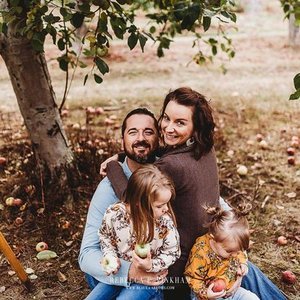 A happy family of four sitting outdoors near a tree, smiling at the camera. The family includes a man, a woman, and two young girls, with the children holding small objects.