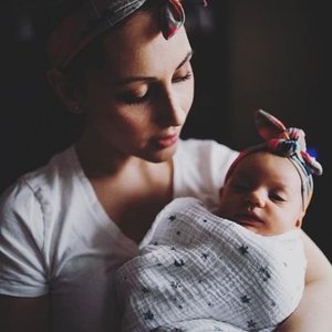 Woman holding a newborn wrapped in a blanket, wearing a headband and sunglasses