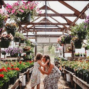 A woman and a young girl in a greenhouse filled with various potted flowers and hanging baskets, with wooden beams overhead, sharing a joyful moment.