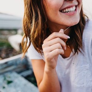 Close-up of a smiling woman with light brown hair holding a toothpick, outdoors in bright natural light.