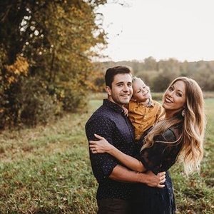 A happy family of three outdoors in a park during autumn, with trees and grass, smiling and embracing.