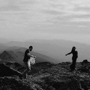 Three people standing on a mountain ridge, overlooking a hilly landscape under a cloudy sky.