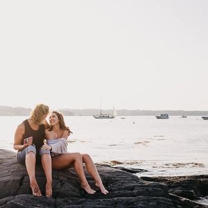Two women sitting on rocks by the water with boats in the background
