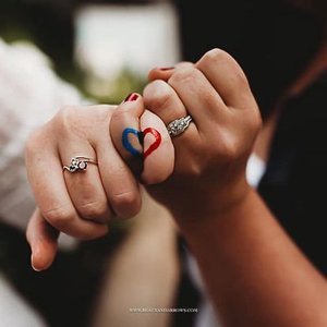 Two people holding hands, showing wedding rings and a small tattoo of a blue and red heart on one finger.