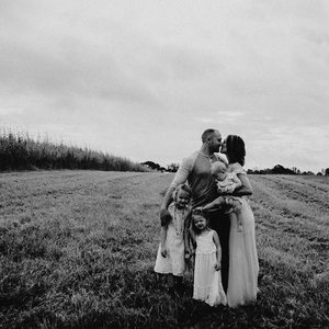 A family of four standing outdoors in a grassy field with overcast sky, embracing each other.