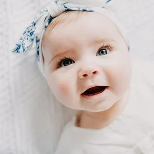 Close-up of a smiling baby girl with light skin, blue eyes, and a blue patterned headband. She is wearing a white shirt and appears to be lying on a white surface.