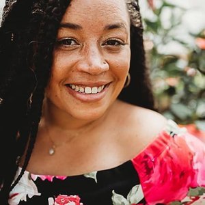A smiling woman with curly hair, wearing a colorful floral off-shoulder top, outdoors with greenery in the background.
