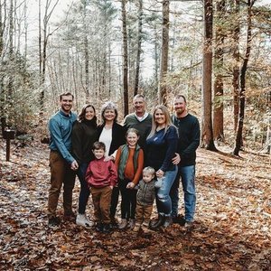 A family of eight standing together in a wooded forest with bare trees, wearing casual fall clothing, smiling for a photo.