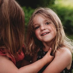 A young girl with long, wavy hair smiling while another child touches her face outdoors.