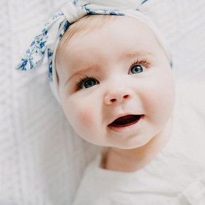 Close-up of a smiling baby girl with a blue patterned headband and light-colored eyes.