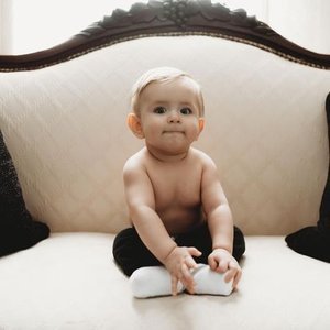 A young toddler sitting on a vintage-style cream-colored sofa, wearing black pants and holding a white sock.