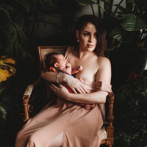 A woman sitting on a chair holding a newborn baby in her arms, surrounded by lush green plants.