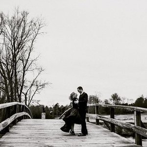 A black and white photo of a couple on a wooden bridge, with one person sitting and the other standing, outdoors with leafless trees in the background.