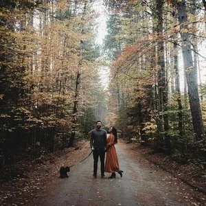 A man, a woman, and two dogs standing on a forest trail during autumn, with trees and fall foliage surrounding them.