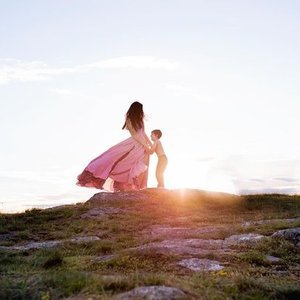 A woman and a child standing on a grassy hill near water at sunset, holding hands.