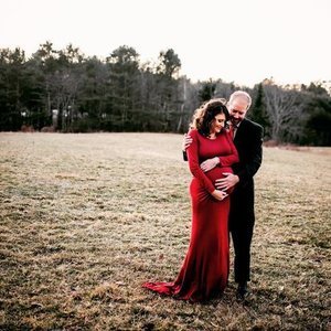 A pregnant woman in a red dress and a man in a dark suit standing outdoors in a field with trees in the background, embracing and smiling.