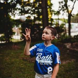 Young boy in a blue sports jersey tossing a baseball in a park with trees in the background.