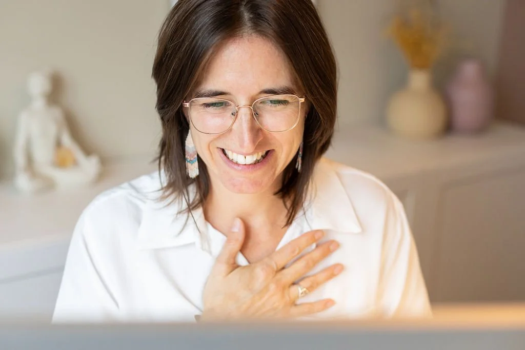 Une femme souriante regarde un écran d'ordinateur, portant des lunettes et une chemise blanche, avec une main sur la poitrine, dans un environnement intérieur décoré avec des vases et une statue en arrière-plan.
