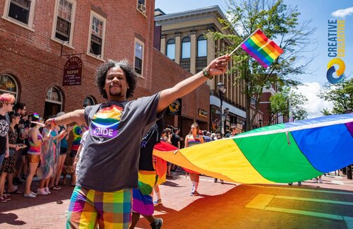 A Black person holds a small rainbow flag on a parade route. They have rainbow checked shorts and a grey t-shirt that says Pride. Behind them other people carry a large rainbow banner horizontal to the ground. Crowds look on behind them.