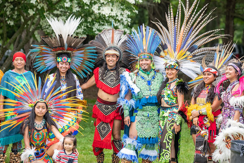 Mashpee Wampanoag tribe members of all ages and genders wear brightly colored traditional costumes with elaborate headdresses. They all smile for the camera.