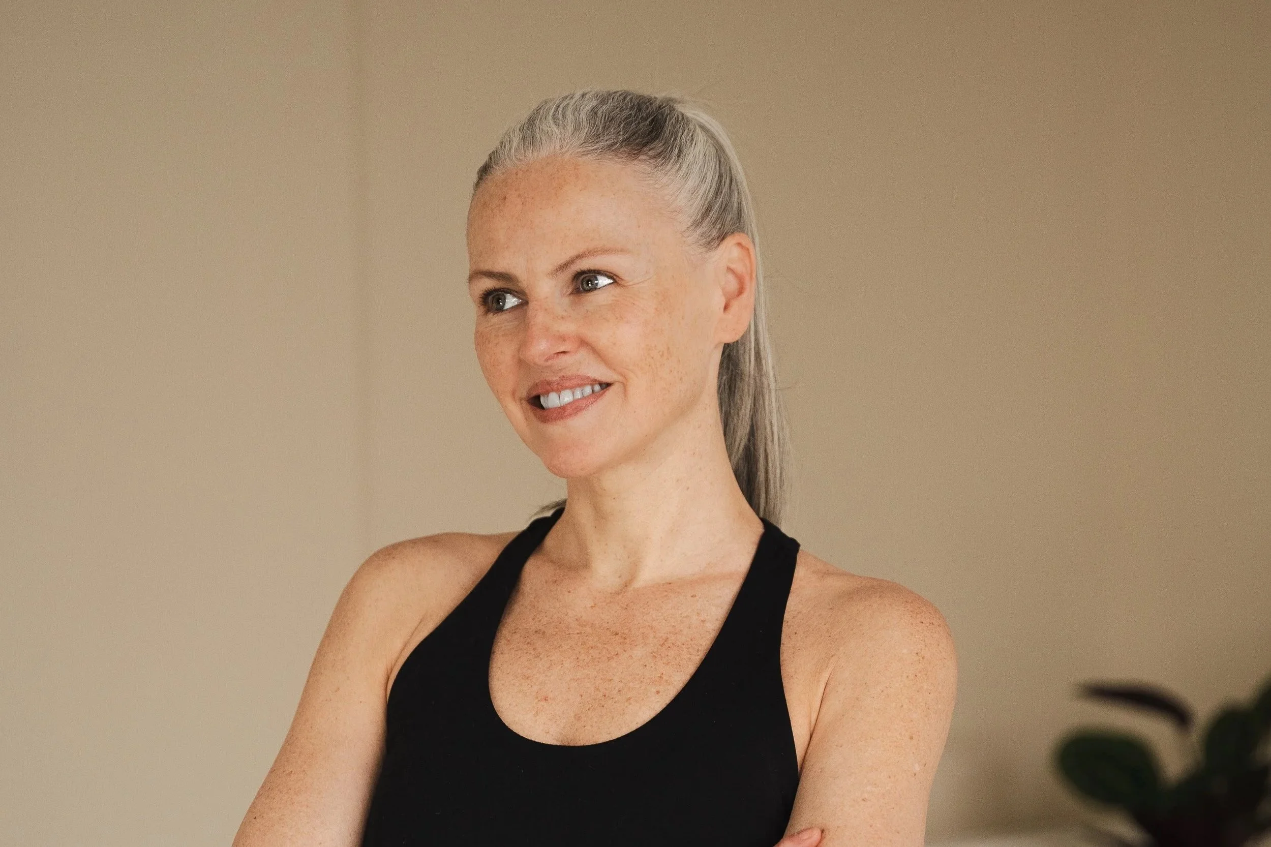 Une femme souriante aux cheveux gris attachés en queue de cheval, portant un débardeur noir, dans une pièce neutre.