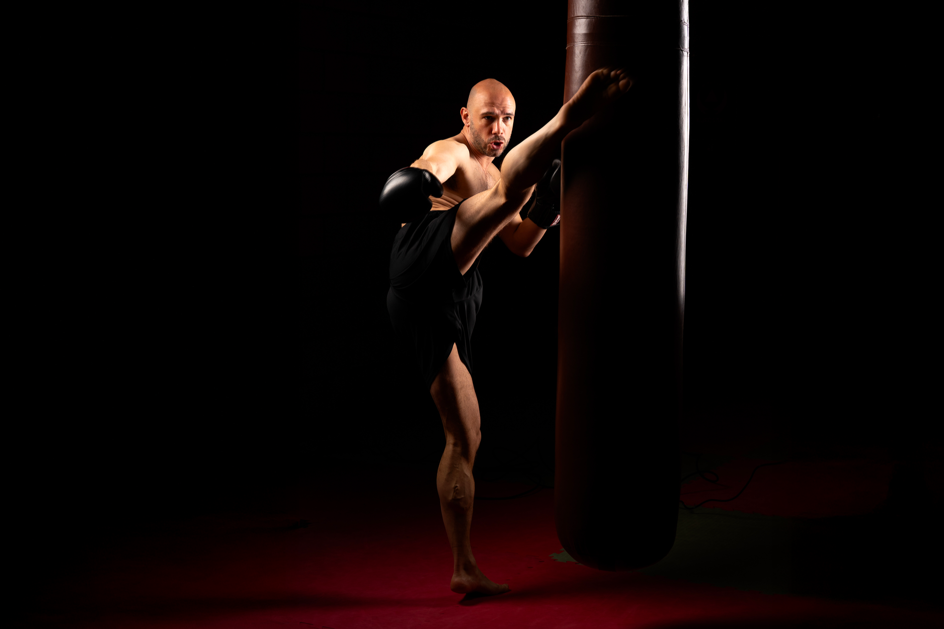 Homme pratiquant un kick à l'entraînement en boxe, en frappant un sac de frappe noir dans une pièce sombre.
