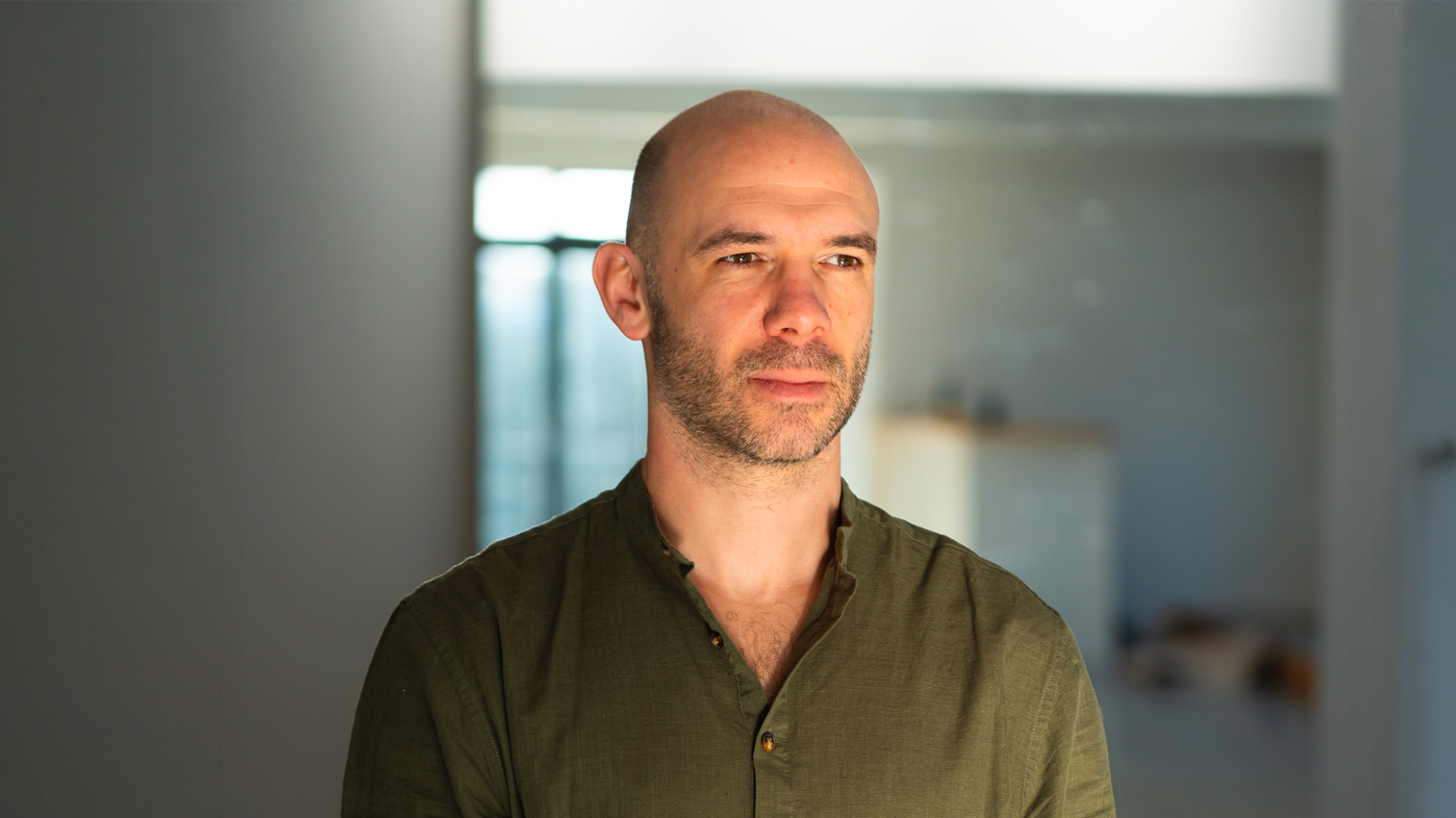 Un homme chauve portant une chemise kaki à col ouvert, se tient dans un intérieur lumineux.