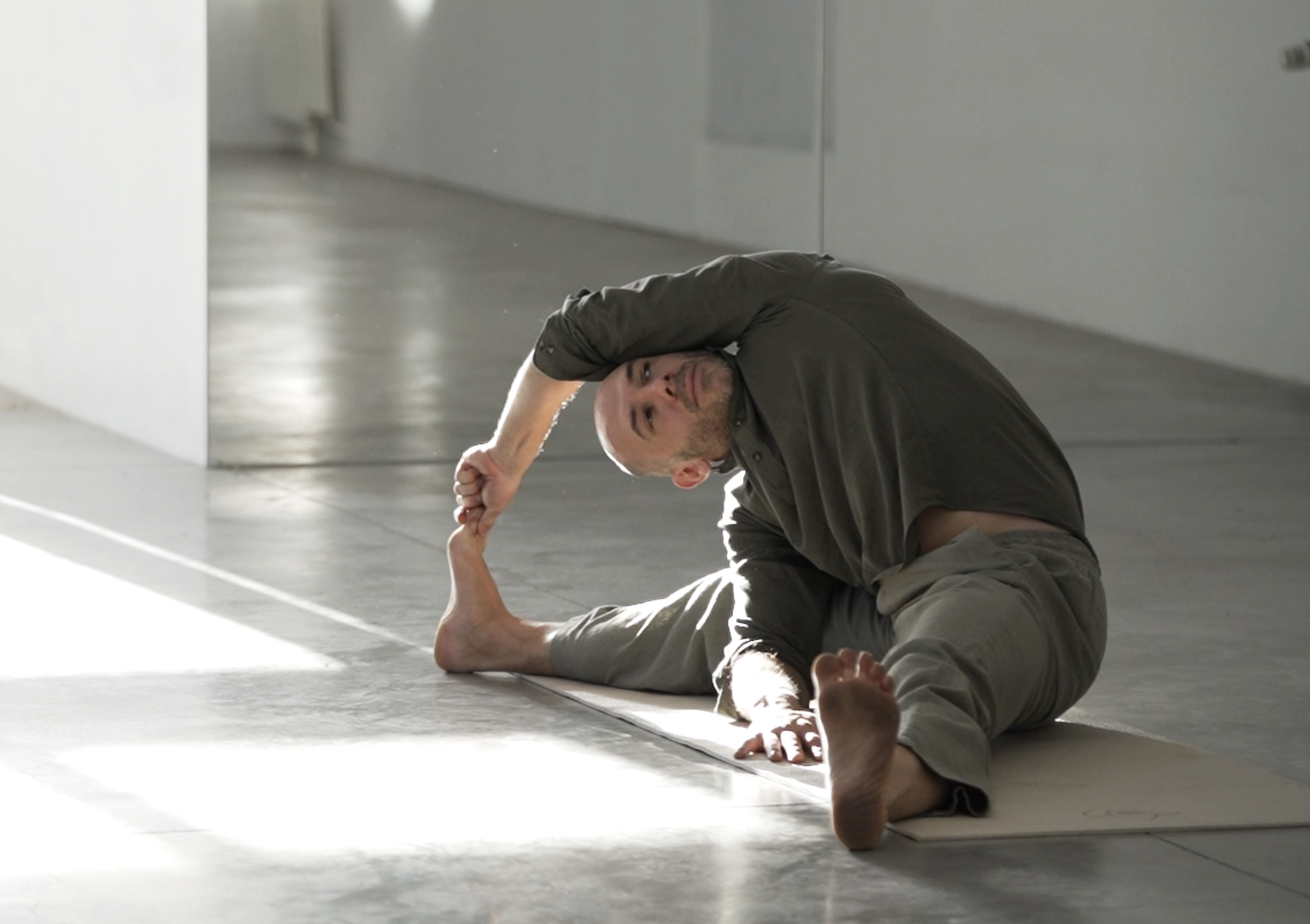 Homme faisant du yoga en position de pince en intérieur, sur un tapis gris, dans un espace épuré avec mur blanc et lumière naturelle.