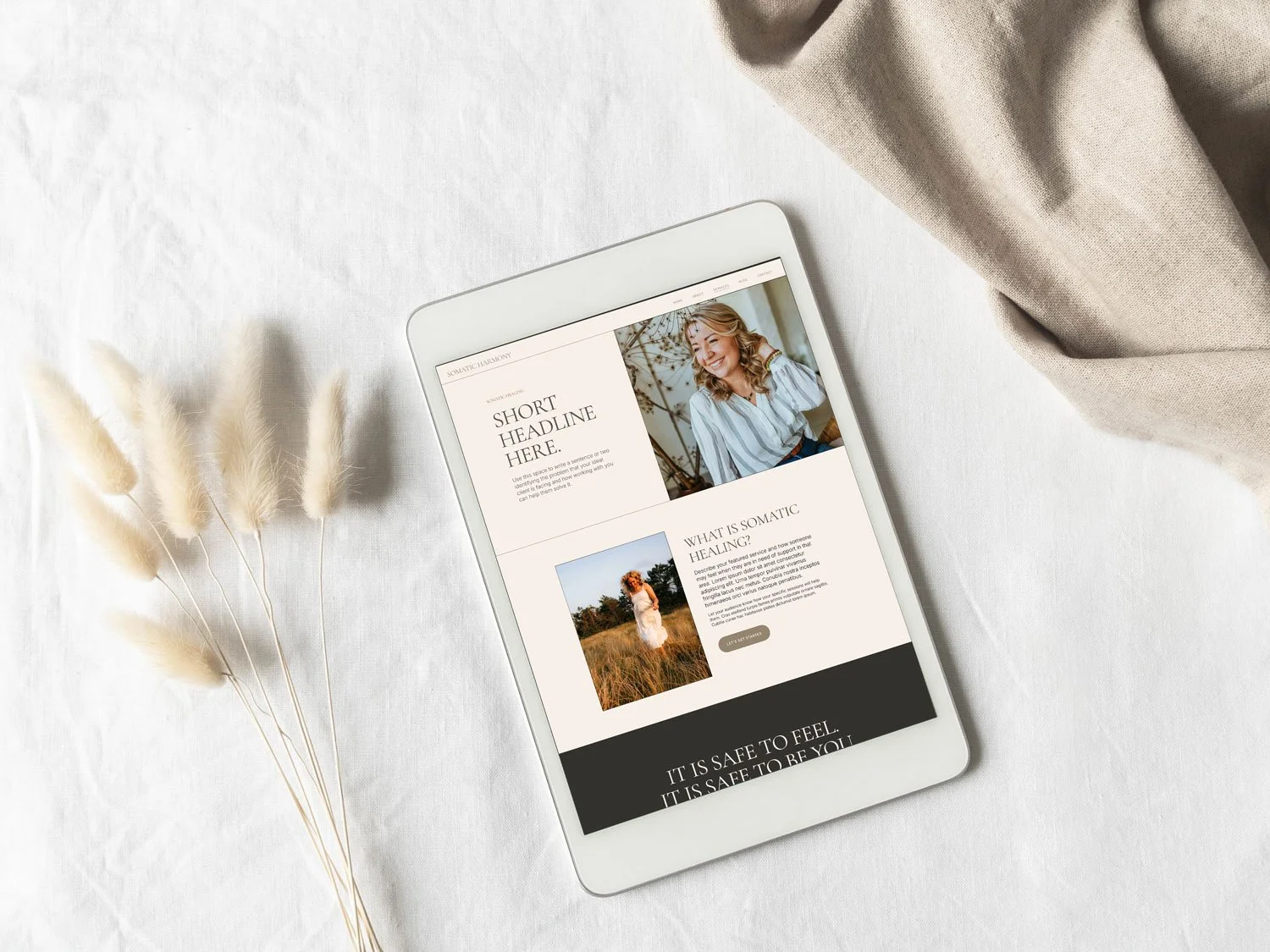A tablet displaying a website about somatic healing, lying on a white fabric surface with a bunch of dried white fluffy flowers nearby and a beige cloth draped in the upper right corner.