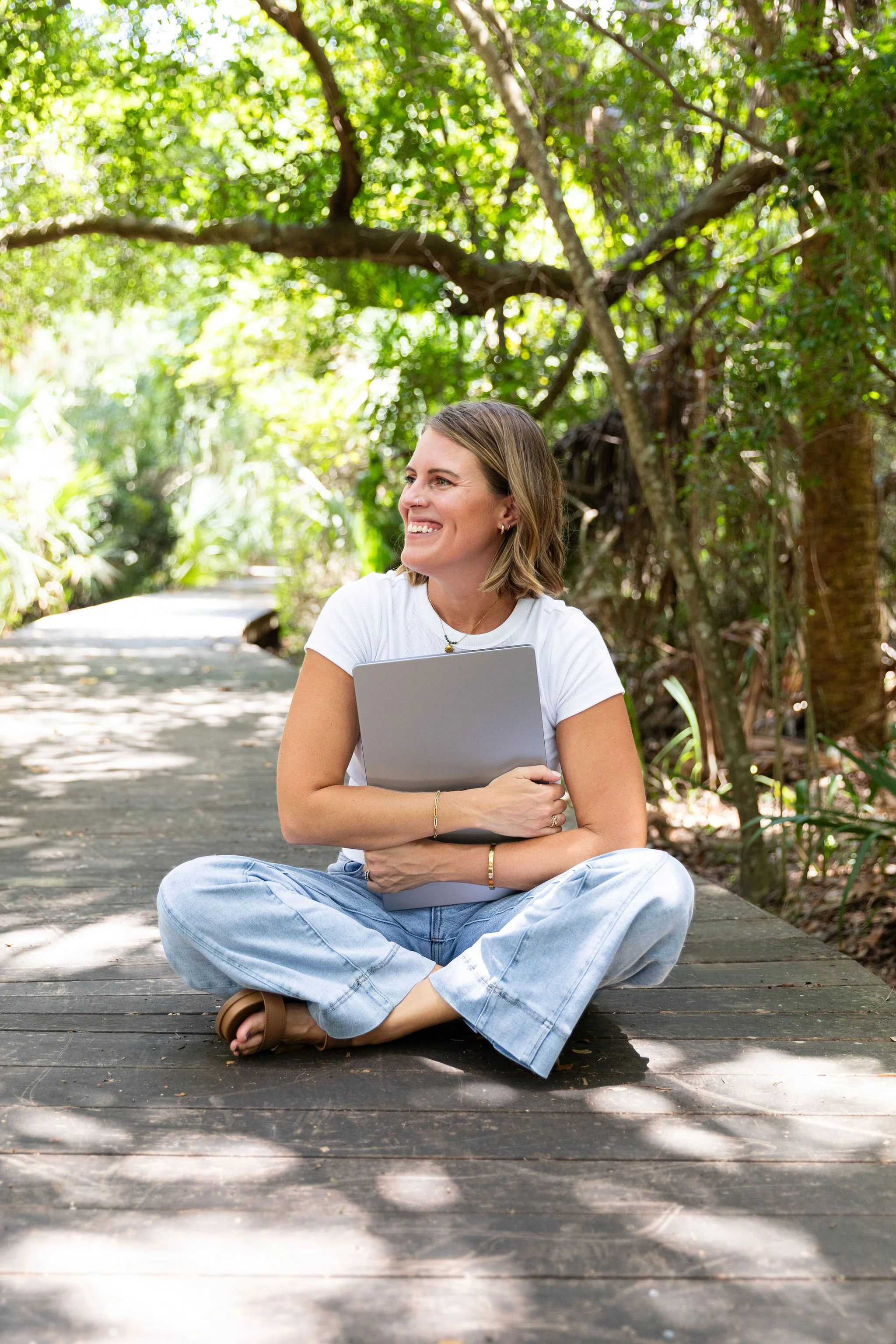 A woman sitting cross-legged on a wooden boardwalk in a lush, green, wooded area holding a closed laptop, smiling and looking to her right.